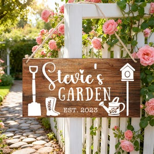 Puede incluir: Un letrero de jardín de madera con el texto "Stevie's GARDEN" y el año de establecimiento "EST. 2023". El letrero presenta ilustraciones blancas de herramientas de jardinería y una casita para pájaros. Cuelga de una valla blanca rodeada de rosas rosas.