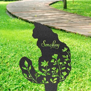 May include: Black metal cat silhouette garden stake with the name "Smokey" in cursive script. The cat's body is filled with a green floral design. The stake is placed in a grassy yard with a wooden walkway in the background.