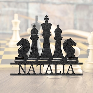 May include: Black metal chess piece decoration featuring a king, two bishops, and two knights. The name "NATALIA" is displayed below the chess pieces. The background shows a blurred chessboard and chess pieces.