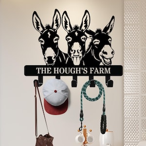 Puede incluir: Perchero de pared de metal negro con tres cabezas de burro y el texto "THE HOUGH'S FARM". Debajo hay ganchos que sostienen una gorra de béisbol, un bolso y una correa para perros. Las cabezas de burro son un elemento decorativo.