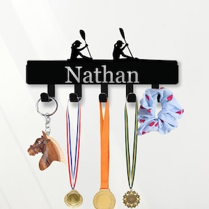 Puede incluir: Soporte de medallas y llaves de metal negro con el nombre "Nathan" en letras plateadas. Presenta siluetas de kayakistas y ganchos que sostienen un llavero de caballo, medallas y una scrunchie azul.