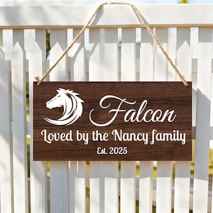 Puede incluir: Un letrero rectangular de madera marrón con texto blanco y un gráfico de cabeza de caballo. El letrero dice "Falcon", "Loved by the Nancy family" y "Est. 2025". Cuelga de una valla blanca con una cuerda de yute.