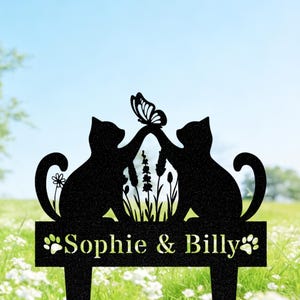 May include: Black metal garden stake with two cat silhouettes and a butterfly. The design includes the names "Sophie & Billy" and paw prints. The background shows a field of white flowers and a blue sky.