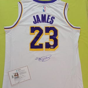 May include: White basketball jersey with purple and gold accents. The jersey features the name "JAMES" and the number "23" on the back. Includes a certificate of authenticity.