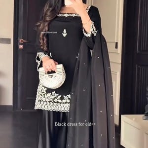 May include: Black dress with white embroidery and a matching black scarf with white dots. The dress has long sleeves and is paired with a white handbag. The text on the image says "Black dress for eid>>".