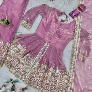 May include: A three-piece ensemble in a soft mauve hue, featuring a tunic, pants, and a scarf. The tunic has a fitted bodice and flared skirt, embellished with intricate gold embroidery. The pants are loose-fitting, and the scarf has a delicate border. A small toy car is on the right.