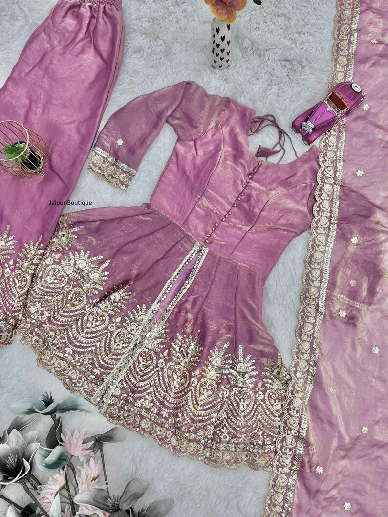 May include: A three-piece ensemble in a soft mauve hue, featuring a tunic, pants, and a scarf. The tunic has a fitted bodice and flared skirt, embellished with intricate gold embroidery. The pants are loose-fitting, and the scarf has a delicate border. A small toy car is on the right.