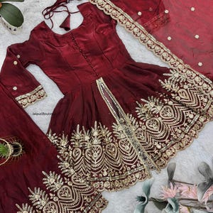 May include: A burgundy-colored, embroidered Indian dress set. The set includes a long-sleeved top with a button-down front, matching pants, and a sheer scarf. The dress is adorned with intricate gold embroidery along the hem and sleeves.