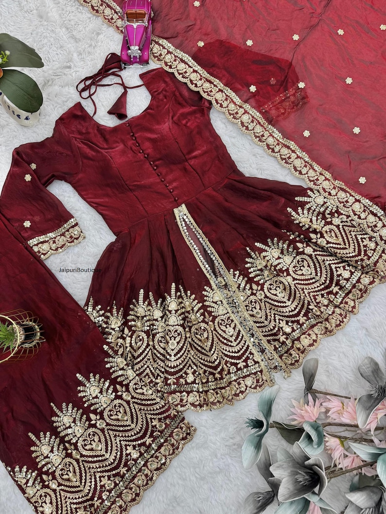 May include: A burgundy-colored, embroidered Indian dress set. The set includes a long-sleeved top with a button-down front, matching pants, and a sheer scarf. The dress is adorned with intricate gold embroidery along the hem and sleeves.