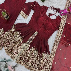 May include: A maroon three-piece outfit featuring a tunic, pants, and a sheer scarf. The tunic and scarf are adorned with gold embroidery. The tunic has a button-down front and three-quarter sleeves. The pants are loose-fitting.