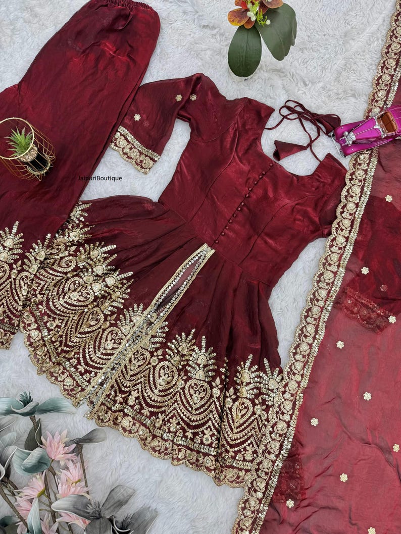 May include: A maroon three-piece outfit featuring a tunic, pants, and a sheer scarf. The tunic and scarf are adorned with gold embroidery. The tunic has a button-down front and three-quarter sleeves. The pants are loose-fitting.