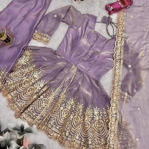 May include: A lavender-colored, three-piece outfit featuring a long-sleeved top with gold embellishments, matching pants, and a sheer scarf. The top has a button-down front and intricate gold embroidery. The pants have a comfortable, elastic waistband.