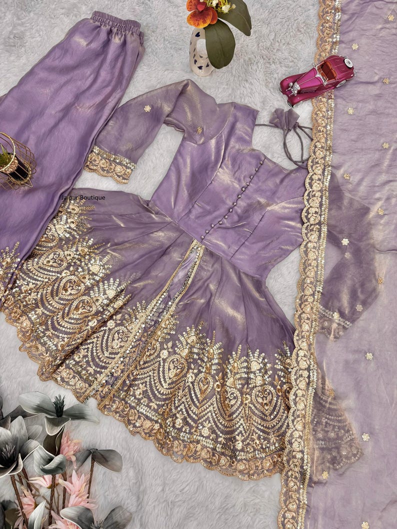 May include: A lavender-colored, three-piece outfit featuring a long-sleeved top with gold embellishments, matching pants, and a sheer scarf. The top has a button-down front and intricate gold embroidery. The pants have a comfortable, elastic waistband.