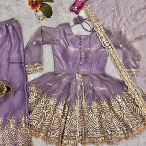 May include: A lavender-colored, three-piece outfit featuring a long-sleeved top with gold embroidery, matching pants, and a sheer scarf. The top has a button-down front and a flared skirt. The outfit is embellished with intricate gold detailing.