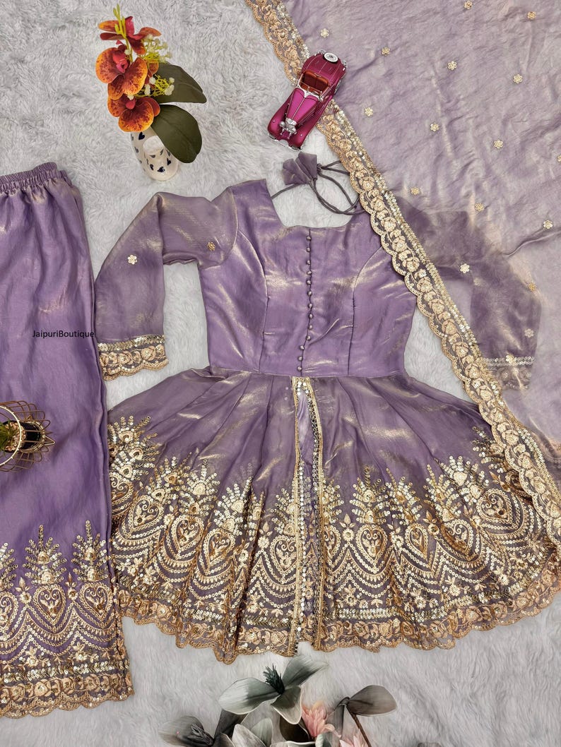 May include: A lavender-colored, three-piece outfit featuring a long-sleeved top with gold embroidery, matching pants, and a sheer scarf. The top has a button-down front and a flared skirt. The outfit is embellished with intricate gold detailing.