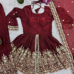 May include: A burgundy-colored, embroidered dress with a matching scarf. The dress features gold embroidery along the hem and sleeves, with a decorative gold trim down the front. The scarf has a gold embroidered border and small, scattered gold embellishments.