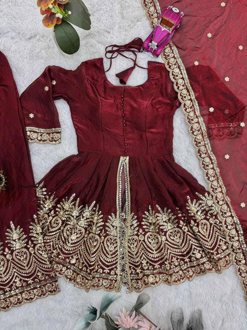 May include: A burgundy-colored, embroidered dress with a matching scarf. The dress features gold embroidery along the hem and sleeves, with a decorative gold trim down the front. The scarf has a gold embroidered border and small, scattered gold embellishments.