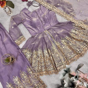 May include: A lavender-colored, long-sleeved dress with intricate gold embroidery along the hem and sleeves. The dress features a fitted bodice and a flared skirt. A matching scarf with gold trim and a pair of lavender pants are also included. A small toy car is visible.
