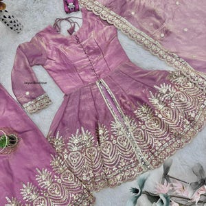 May include: A lilac-colored, embroidered formal outfit. The set includes a long-sleeved top with a button-down front, matching pants, and a sheer scarf. The outfit features intricate gold embroidery along the edges and hem.
