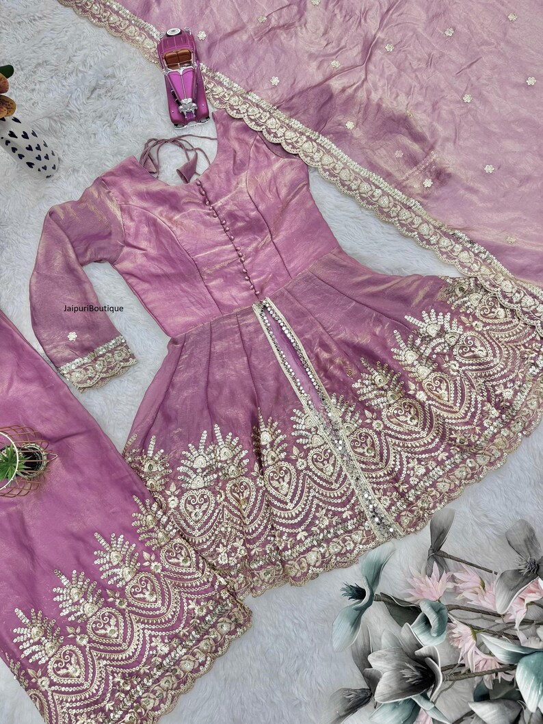 May include: A lilac-colored, embroidered formal outfit. The set includes a long-sleeved top with a button-down front, matching pants, and a sheer scarf. The outfit features intricate gold embroidery along the edges and hem.