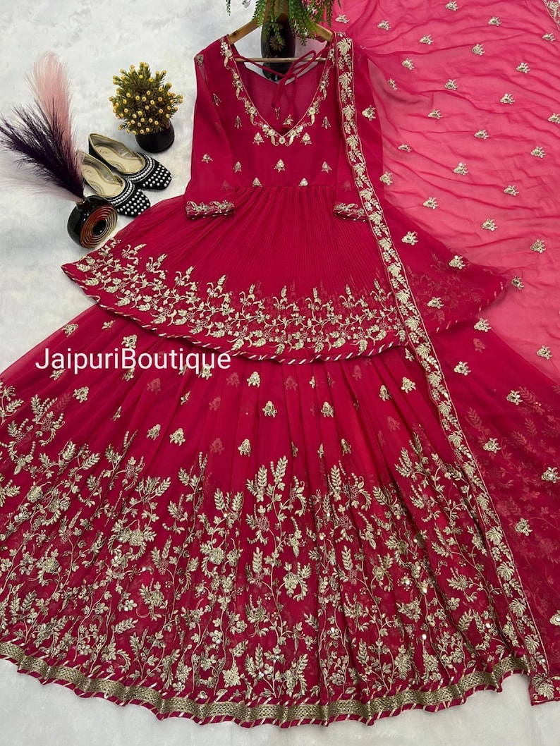 May include: A vibrant fuchsia-colored ensemble featuring intricate gold embroidery. The outfit includes a long-sleeved top with a V-neck and a tiered skirt. The text "Jaipuri Boutique" is visible. A matching dupatta completes the look.