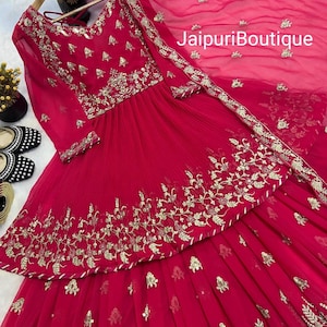 May include: A vibrant red ensemble featuring intricate gold embroidery. The outfit includes a long-sleeved top and a matching skirt, with a sheer dupatta. The text "JaipuriBoutique" is visible on the dupatta.