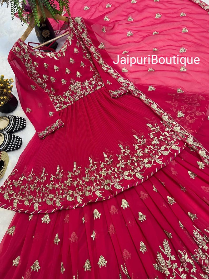 May include: A vibrant red ensemble featuring intricate gold embroidery. The outfit includes a long-sleeved top and a matching skirt, with a sheer dupatta. The text "JaipuriBoutique" is visible on the dupatta.
