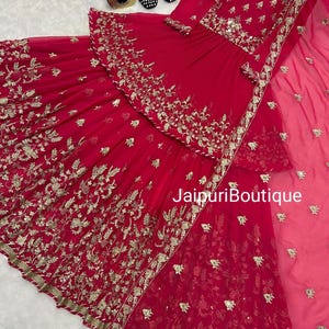 May include: A vibrant red ensemble featuring intricate gold embroidery. The outfit includes a top and a flowing skirt, with a matching sheer scarf. The text "JaipuriBoutique" is visible on the garment.