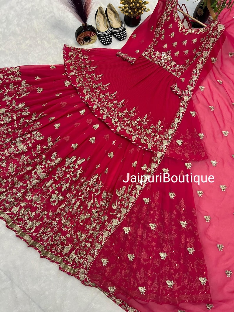 May include: A vibrant red ensemble featuring intricate gold embroidery. The outfit includes a top and a flowing skirt, with a matching sheer scarf. The text "JaipuriBoutique" is visible on the garment.