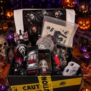 Op de afbeelding: Een zwarte geschenkdoos omwikkeld met geel waarschuwingslint met horror-merchandise, waaronder een beker met een Ghostface-afbeelding, een Michael Myers Tiny Nightmares-figuurtje, een stoffen tas met kettingzaagprint en een strik met patroon.