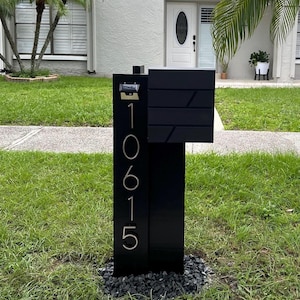 May include: A modern black mailbox with the address numbers "10615" in gold. The mailbox has a rectangular design with a slot for mail and a separate compartment. It is set on a bed of black stones.