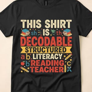 May include: A black short-sleeved t-shirt featuring a graphic design with the text "THIS SHIRT IS DECODABLE STRUCTURED LITERACY READING TEACHER". The design includes colourful illustrations of books, a graduation cap, pencils, an apple, and stars.