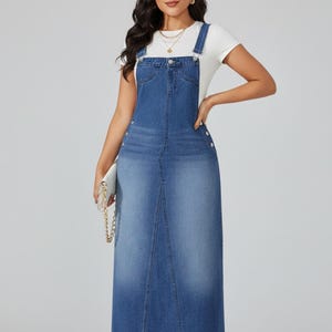 Puede incluir: Un vestido largo con peto de mezclilla en un lavado azul medio. El vestido presenta un frente de peto con bolsillos, tirantes ajustables y cierres de botones en los laterales. El vestido se usa sobre una camiseta blanca.