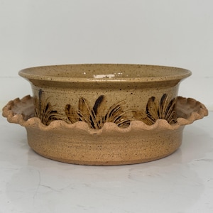 May include: A tan ceramic bowl with a matching base. The bowl has a slightly flared rim and is decorated with a brown floral pattern. The base has a scalloped edge. The bowl and base are speckled.