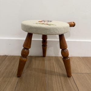May include: A small, three-legged wooden stool with a round, beige seat. The seat features a floral needlepoint design in various colors. The legs and handle are made of polished, dark brown wood.