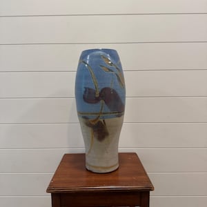May include: A tall, ceramic vase with a wide base and a narrow neck. The vase features a blue upper section with brown and gold leaf-like designs. The lower section is a light beige color. The vase is displayed on a small, dark wooden table.