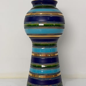 May include: A tall, ceramic vase with a striped design. The vase features bands of navy blue, turquoise, and green, separated by gold-coloured stripes. The top and base have a gold trim. The vase has a glossy finish.