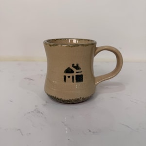 Op de afbeelding: Tan keramische mok met een zwart huisje. De mok heeft een gebogen vorm en een afgeronde handgreep. De rand en de basis hebben een donkerdere, getextureerde rand. De mok is waarschijnlijk handgemaakt.