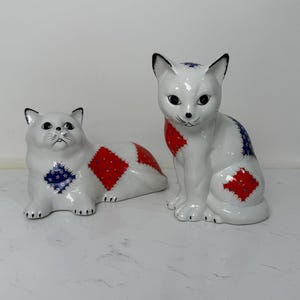 May include: Two white ceramic cat figurines. One is lying down, the other is sitting upright. Both have black accents and patches of red and blue patterned fabric. The cats have detailed facial features.