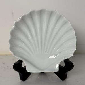 May include: A white, seashell-shaped serving dish. The dish has a scalloped edge and a ribbed interior, resembling a natural scallop shell. It is displayed on a black stand, perfect for serving appetizers or displaying decorative items.