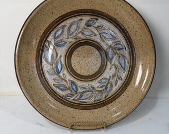 May include: A large, round ceramic plate with a speckled tan rim and a central design. The center features a ring of blue and white leaves around a brown circle. The plate is displayed on a gold-colored stand.
