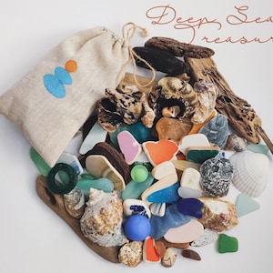 May include: A collection of sea treasures, including seashells, sea glass, and driftwood, arranged on a white surface. A beige drawstring bag with a blue and orange design is on the left. The words "Deep Sea treasure" are written in cursive.