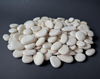 Puede incluir: Un montón de piedras de río blancas y lisas de varios tamaños. Las piedras tienen una forma natural y redondeada y una superficie ligeramente texturizada. Las piedras están dispuestas sobre un fondo gris oscuro, creando un contraste.