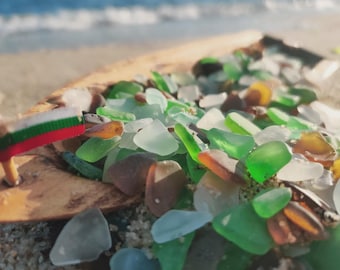 May include: Close-up of sea glass in shades of green, white, and amber, resting on a piece of driftwood on a sandy beach. A small flag with red, white, and green stripes is visible. The ocean is blurred in the background.