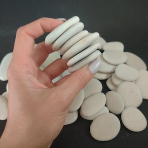 Puede incluir: Una mano sosteniendo una pila de piedras de río lisas y de color gris claro, con un montón de piedras similares en el fondo. Las piedras son ovaladas y planas, con un aspecto natural y desgastado. El fondo es oscuro.