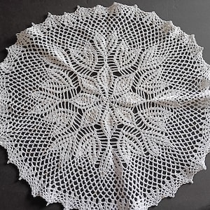 May include: A white, circular, crocheted doily with a detailed floral design in the centre. The doily has a scalloped edge and is made of a fine, open-weave material. The intricate pattern is visible throughout the piece.