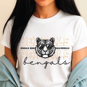 Puede incluir: Camiseta blanca con un tigre en blanco y negro con gafas de sol. La camiseta tiene la palabra "BENGALS" en una fuente estilizada, con la palabra "bengals" en cursiva debajo del gráfico del tigre.