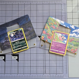 Peut inclure: Deux cartes à collectionner Pokémon, l'une avec Noctali et l'autre Rondoudou, sont présentées dans des pochettes de protection transparentes. La carte Noctali a un fond sombre avec une lune, tandis que la carte Rondoudou a un personnage rose. Les cartes sont sur une surface quadrillée.
