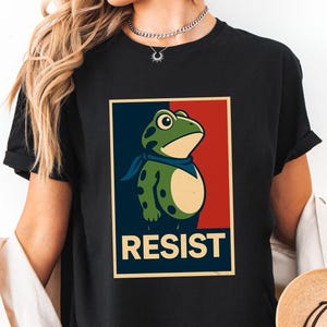 May include: Black t-shirt featuring a cartoon frog with a blue bandana, set against a red and blue background. The word "RESIST" is printed below the frog in bold white letters. The shirt is a casual wear item.