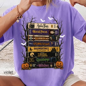 Op de afbeelding: Een violet t-shirt met een grafische print van een stapel van acht vintage VHS-banden met de titels The Addams Family, Hocus Pocus, Beetlejuice, Halloweentown, Halloweentown II, The Nightmare Before Christmas, Goosebumps en The Witches.